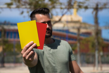 Handsome young man holding a football in his hand while showing red and yellow card to camera as if...