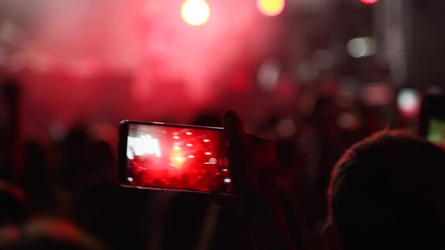 People silhouettes waving hands, taking photos or recording videos of live music concert with smartphones at open air festival. Bright colorful stage lighting. Fans at favourite band performance.