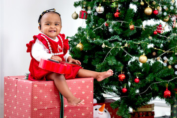 Portrait of adorable happy smiling African American little girl child sitting on big gift box present under Christmas tree in living room, kid celebrating happy Christmas winter holiday