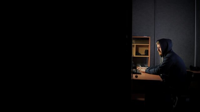 A bearded man in a hood works at a computer by the light of a desk lamp. Male office worker at night in a dark room. Work online with documents. Hacker or programmer
