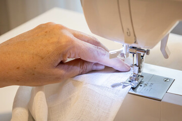 Cropped female hands sewing fabric in manufacturing machine at workplace. Needle pressure foot with blurred white background. Apparel production. Close up top side view. Selective focus copy space.