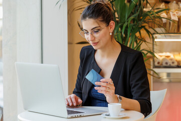 una ragazza bianca seduta in un tavolino dal proprio portatile  , con una carta di credito in mano  è pronta a fare degli acquisti vi internet