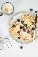 Homemade viennese waffles with blueberries and banana and cup of coffee on a light background.