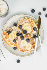 Homemade viennese waffles with blueberries and banana and cup of coffee on a light background.