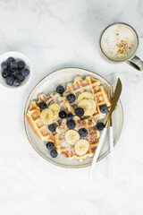 Homemade viennese waffles with blueberries and banana and cup of coffee on marble background.