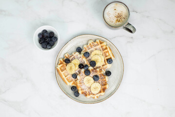 Homemade viennese waffles with blueberries and banana and cup of coffee on marble background.
