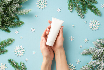 Winter season skin care concept. First person top view photo of young woman's hands holding white tube without label over snowflakes and pine branches in frost on isolated pastel blue background