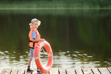 A boy in a life jacket and with a life buoy is standing near the water in the rays of the setting...