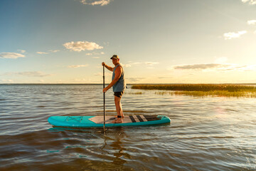 Plakat a man in shorts with a paddle on a sup board at sunset in the lake.