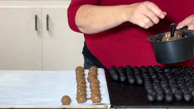 4K HD video of an older caucasian female in kitchen dipping cake balls in chocolate candy melted, then dropping coated cake balls with a fork on a cookie sheet to set up.
