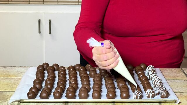 4K HD video of an older caucasian female in kitchen accenting cake balls with white candy melted, striping the topping onto chocolate dipped cake balls on a cookie sheet.
