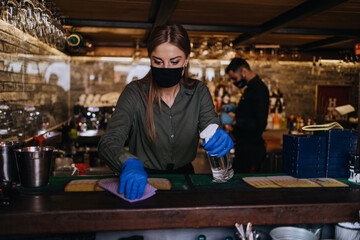 Fototapeta na wymiar Portrait of a serious waitress standing behind a bar in a nice restaurant. She wears a protective mask and gloves as part of security measures against the Coronavirus pandemic.