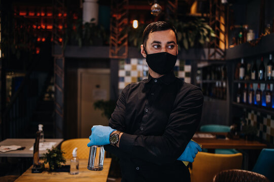 Portrait of a serious waiter standing in a nice restaurant. He wears a protective mask and gloves as part of security measures against the Coronavirus pandemic.