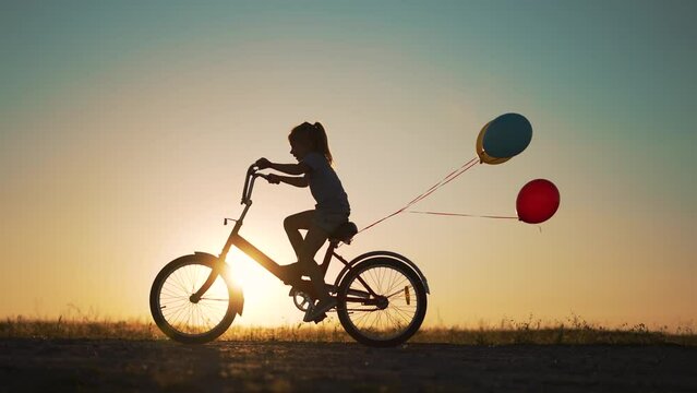 Dream kid. Silhouette of kid on bike in park. Girl rides in park on green grass. Child games in nature.Traveling with balloons on bike.Active child freedom in summer.Girl learns to ride bike in nature