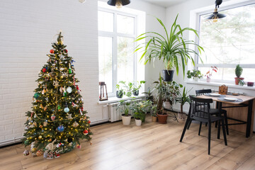 Christmas tree in the white interior of a house with large windows. Glowing fairy lights garlands interior decoration of the studio room. Potted plants in the home