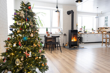 Festive interior of house is decorated for Christmas and New Year in loft style with black stove, fireplace, Christmas tree. Warm studio room with white kitchen, burning wood, cozy and heating of home