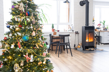 Festive interior of house is decorated for Christmas and New Year in loft style with black stove, fireplace, Christmas tree. Warm studio room with white kitchen, burning wood, cozy and heating of home