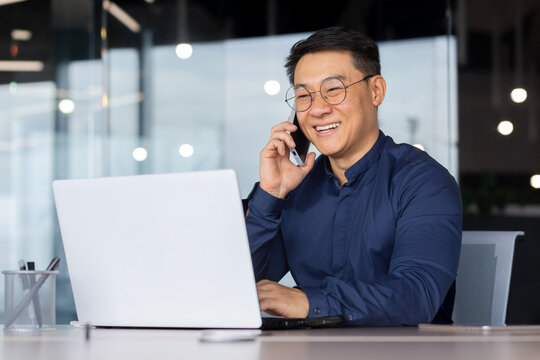 Successful Asian Man Smiling And Talking On The Phone, Businessman In A Shirt Working Inside The Office Using A Laptop.