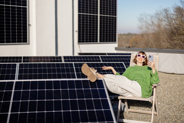 Young woman talks on phone while sitting relaxed on rooftop with a solar station. Happy owner of...