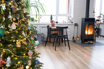 Fototapeta na wymiar Festive interior of house is decorated for Christmas and New Year in loft style with black stove, fireplace, Christmas tree. Warm studio room with white kitchen, burning wood, cozy and heating of home