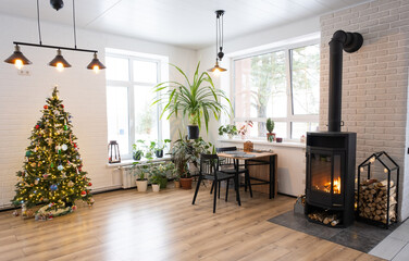 Festive interior of house is decorated for Christmas and New Year in loft style with black stove, fireplace, Christmas tree. Warm studio room with white kitchen, burning wood, cozy and heating of home