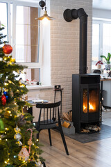 Festive interior of house is decorated for Christmas and New Year in loft style with black stove, fireplace, Christmas tree. Warm studio room with white kitchen, burning wood, cozy and heating of home