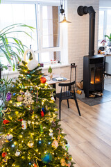 Festive interior of house is decorated for Christmas and New Year in loft style with black stove, fireplace, Christmas tree. Warm studio room with white kitchen, burning wood, cozy and heating of home