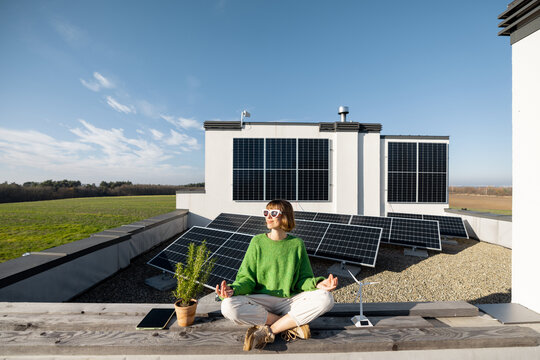 Young woman meditates on rooftop of her house with solar power plant on background. Concept of energy balance and independence