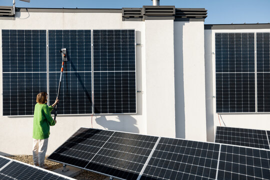 Woman washing solar panels with a brush on rooftop of her household on sunny day. Solar station care and maintenance concept