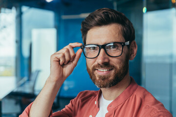 Closeup photo of young successful architect with beard, mature male designer looking at camera and smiling in glasses and red shirt near window inside office.