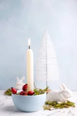 Foto auf Leinwand White burning candle, moss, briar berries,  white rabbits and white decorative tree on white marble background against blue  textured wall. Rabbit is symbool of 2023.. Selective focus. Place for text. © daffodilred
