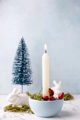 Foto auf Leinwand White burning candle, moss, briar berries,  white bunnies and blue decorative tree on white marble background against blue  textured wall. Rabbit is symbool of 2023.. Selective focus. Place for text. © daffodilred