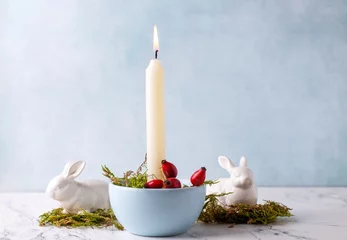 Foto auf Leinwand White burning candle, moss, briar berries and white bunnies on white marble background against blue  textured wall. Rabbit is symbool of 2023.. Selective focus. Place for text. © daffodilred
