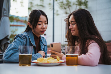 Two young women looking at photos on a smart phone while they are sitting at bar terrace