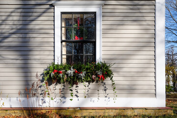 A garland of green ivy, fir tree branches, red ribbon, silver Christmas decorations, and red berries hanging under a vintage casement glass window. The exterior wall is a tan in color with white trim 