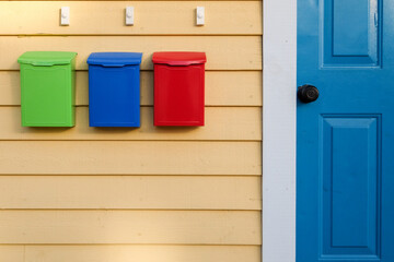 Colorful mailboxes of green, blue, and red on a yellow wooden clapboard exterior wall of a house with a bright blue door. There a three white doorbell buttons over the brightly colored postal boxes. 