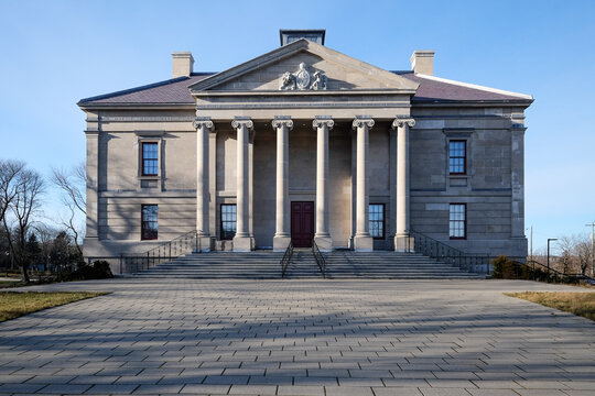 The exterior facade of a historic government building with large vintage marble pillars or columns, red door, blue sky, trees, and a brick entrance leading up steps to the large antique stone building