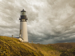 A white high lighthouse on the green shore of the ocean against the background of a gloomy sky with...