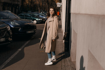 Fototapeta premium Young woman in a coat outside. Outdoor autumn portrait of young elegant fashionable woman wearing trendy glasses, camel color coat, shoulder bag, walking in street of European city. Copy space