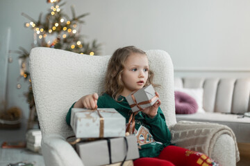 Obraz na płótnie Canvas Beautiful baby child girl in fashionable Christmas clothes sits in an vintage chair with a gifts on the background of a Christmas tree at home on Christmas Eve