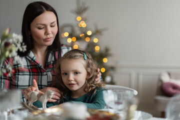 Beautiful mother and daughter in fashionable clothes are sitting at the festive table against the background of the Christmas tree on Christmas Eve