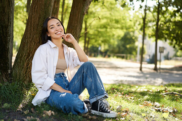 Stylish young university student, girl sits in park on lawn, leans on tree and smiles, resting outdoors and enjoying nature