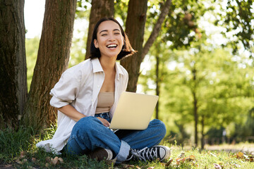 Portrait of asian girl with laptop, sitting near tree in park on sunny summer day, working on remote, e-learning with computer