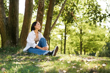Portrait of asian girl relaxing, leaning on tree and resting in park under shade, smiling and enjoying the walk outdoors
