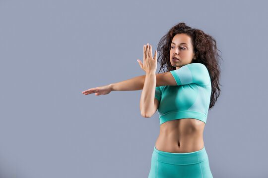 Fit girl doing cross-body shoulder stretch studio. Active athlete stretching isolated on grey.