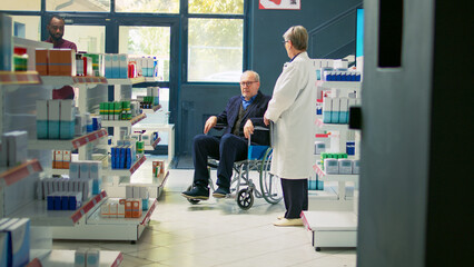 Pharmacist giving cardiology pills bottle to senior wheelchair user, man with chronic disability asking for support. Client with impairment receiving pharmaceutics treatment in pharmacy.