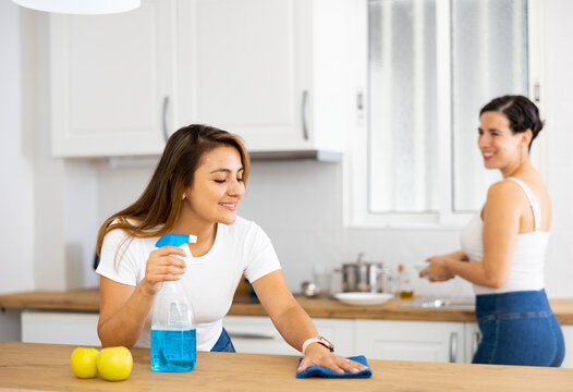 Positive Interested Young Latin American Woman With Sister Cleaning House During Day, Wiping Kitchen Countertop With Rag And Spray