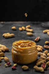 Peanut butter or paste in an open jar and peanuts in the peel scattered on dark background, American dessert concept. vertical image. top view. place for text