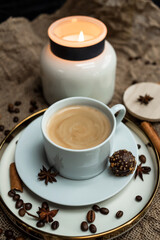 An aesthetic composition of a cup of black coffee with foam and a burning candle, decorated with chocolates, anise stars, coffee beans and cinnamon sticks