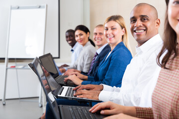 Side view of cheerful hispanic businessman working on laptop with group of colleagues during corporate seminar